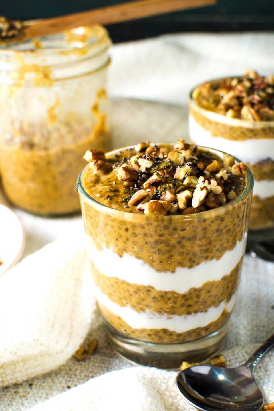 Two glasses filled with 5 layers of chia pudding and cream, with nuts on top and a mostly empty glass in the background.