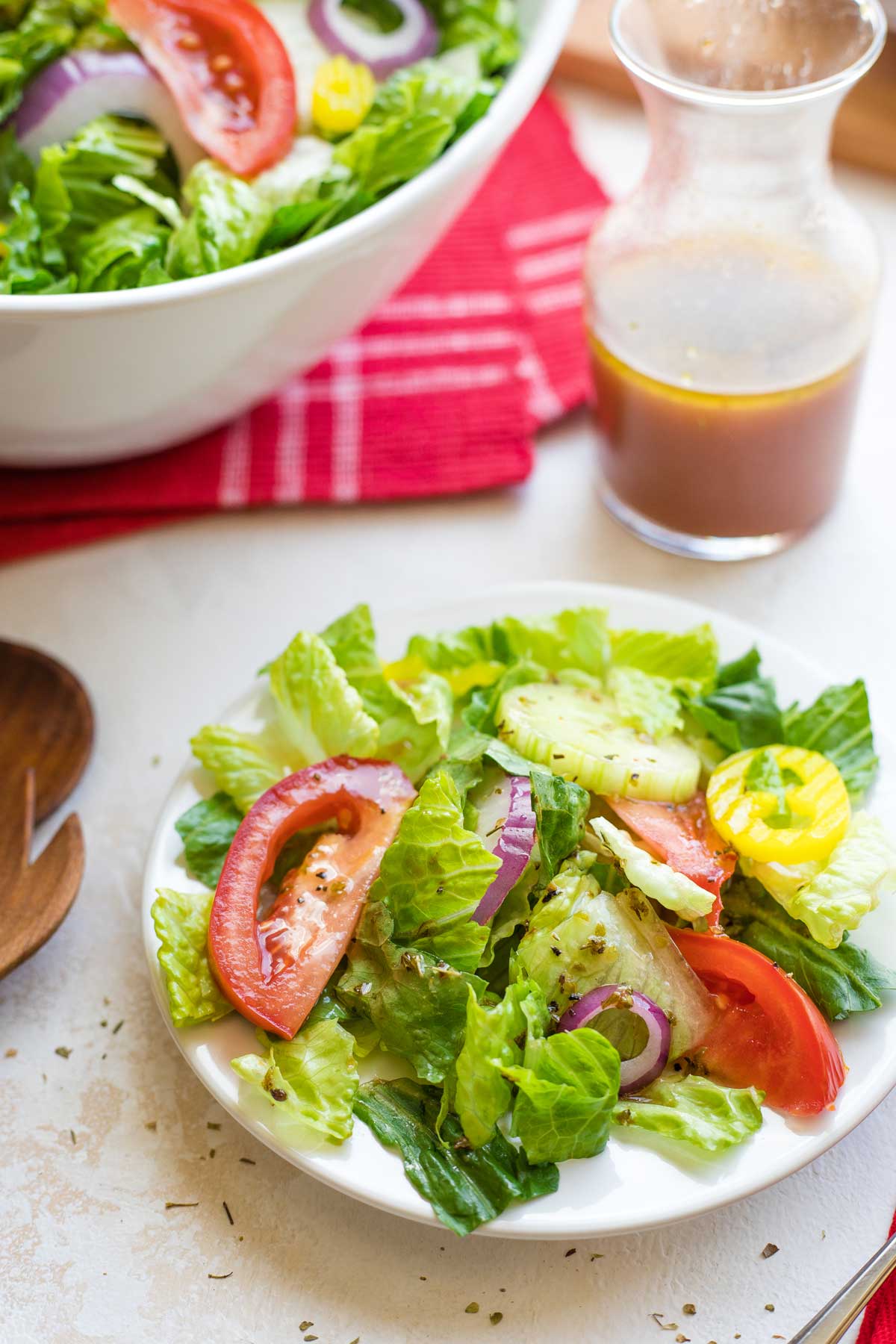 Small plate of Italian salad with bottle of vinaigrette and serving bowl of salad on red cloth behind.