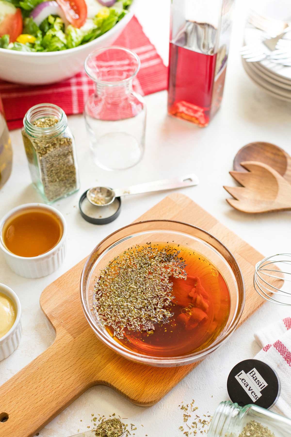 Clear glass bowl of unmixed dressing with spices, bowls of honey and mustard, bottle of red wine vinegar, and bowl of salad.