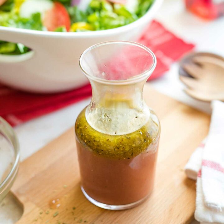 Cruet of vinaigrette on wooden board with bowl of Italian salad on red cloth, red wine vinegar and salad servers.