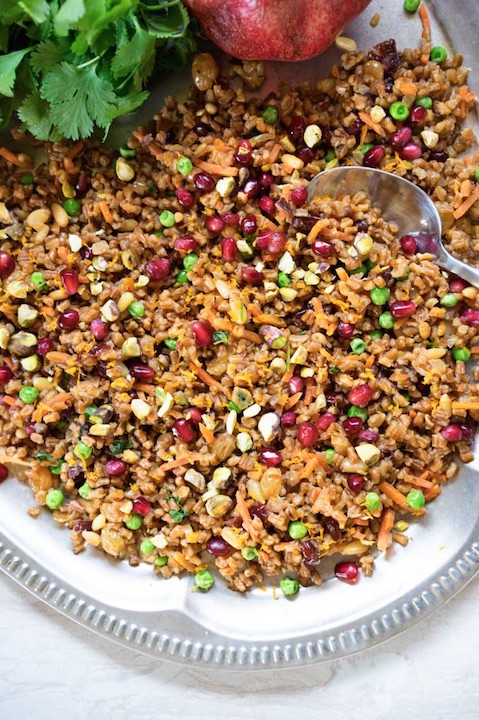 Overhead of grain salad on gray-rimmed, white-scalloped plate with spoon, herbs and whole pomegranate at edges.