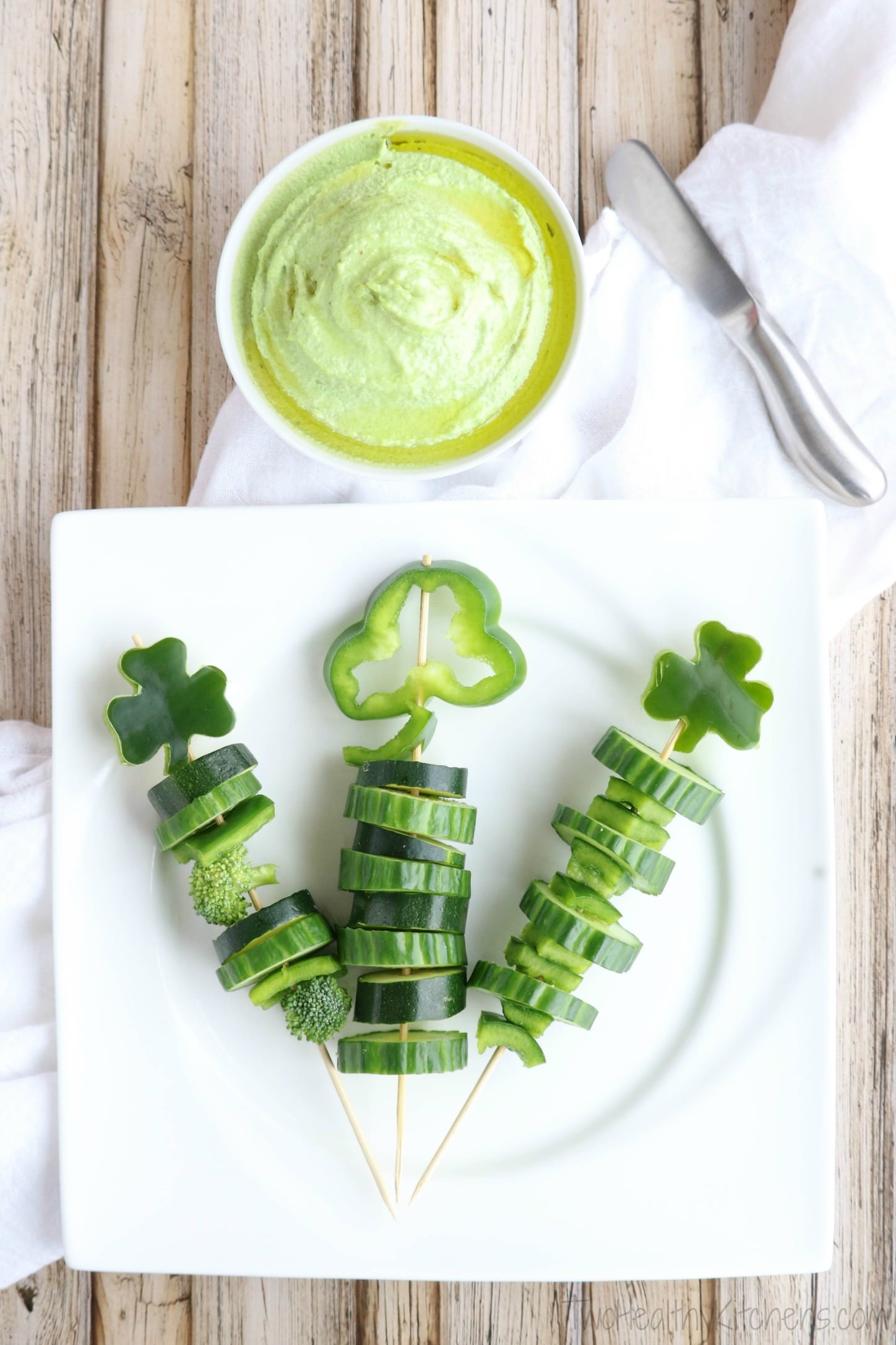 Vertical image of skewers arrayed on plate with various arrangements of green veggies with white cloth, bowl of green dip and dip spreader.