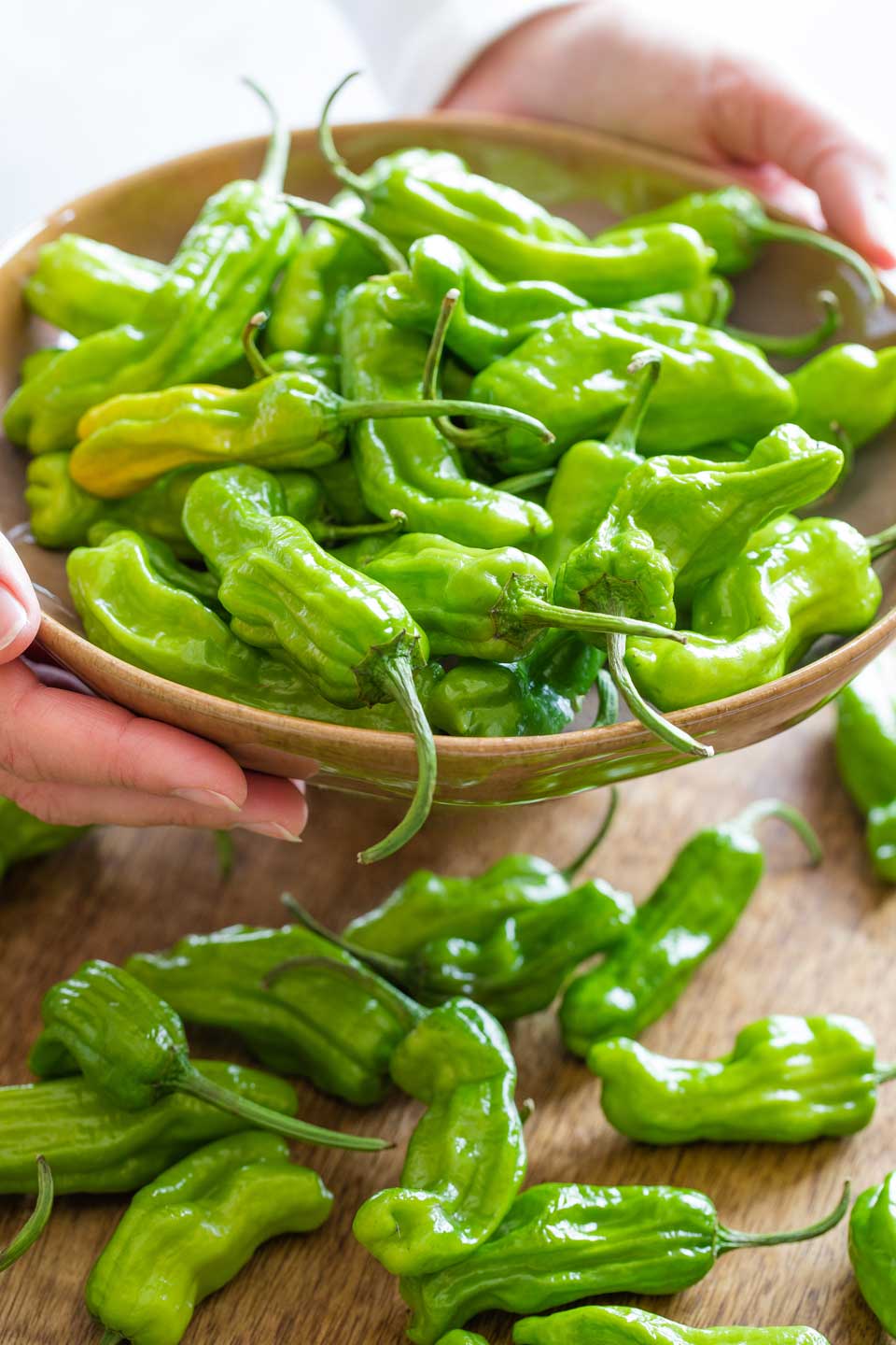 Two hands holding a bowl full of raw shishitos, with other peppers scattered on a board beneath.