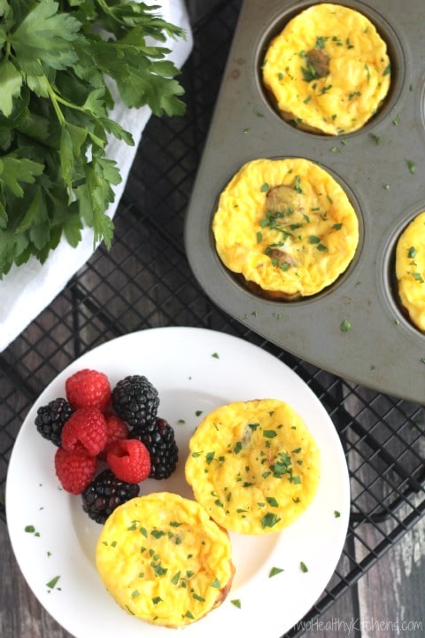 Flatlay scene with 2 egg cups and berries on small white plate on baking rack; muffin tin with more egg ups and fresh parsley surrounding.