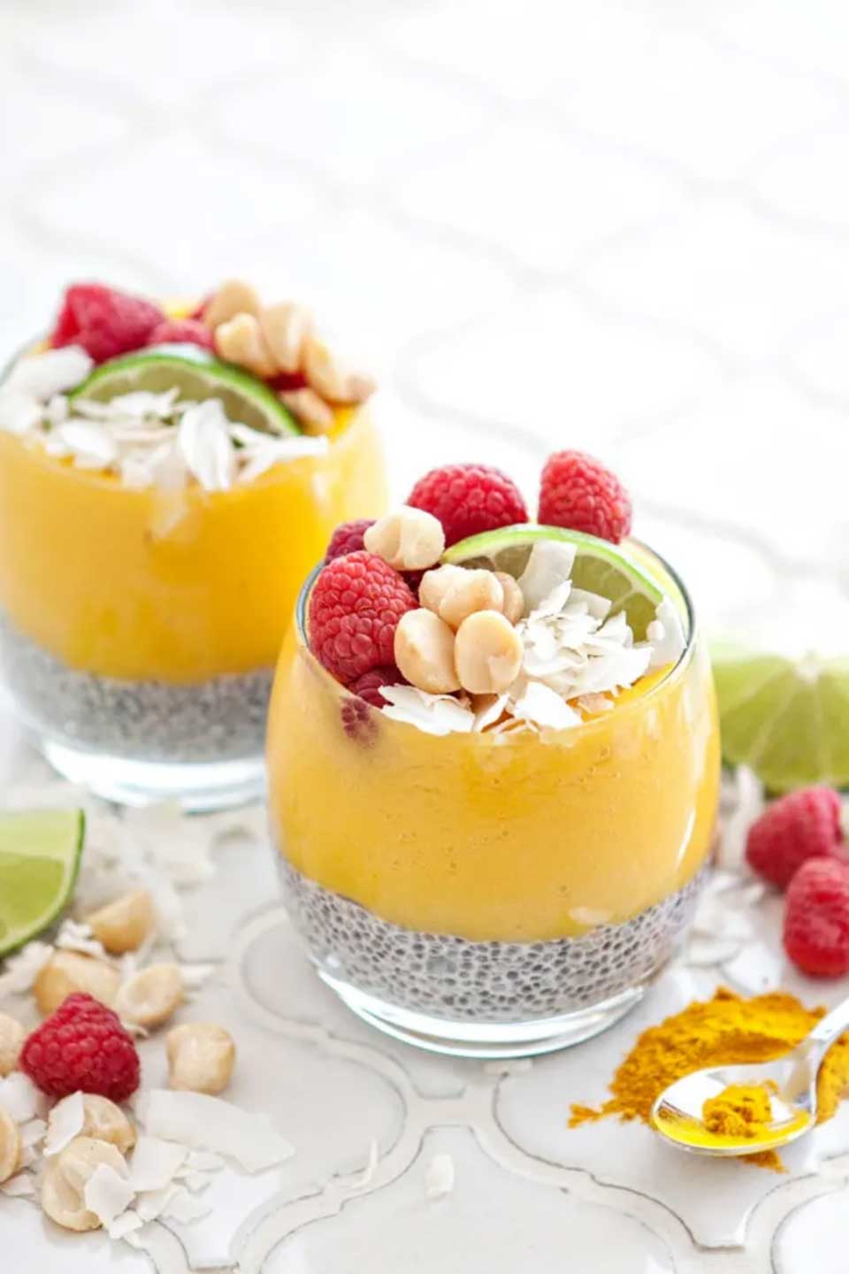 Two glasses layered with chia pudding and fruit puree, garnished with raspberres, coconut, macadamias and lime wedges.