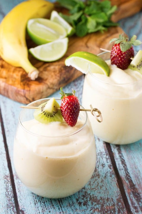 Two glasses of smoothie topped with little skewer of fruit, with banana, lime wedges and herbs on cutting board in background.