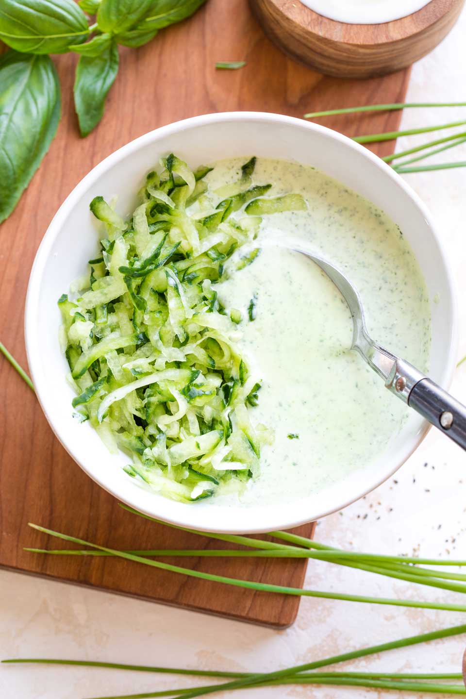 Overhead of a white bowl full of the blended tzatziki sauce, with a pile of cucumber shreds on the left side, ready to be stirred in.