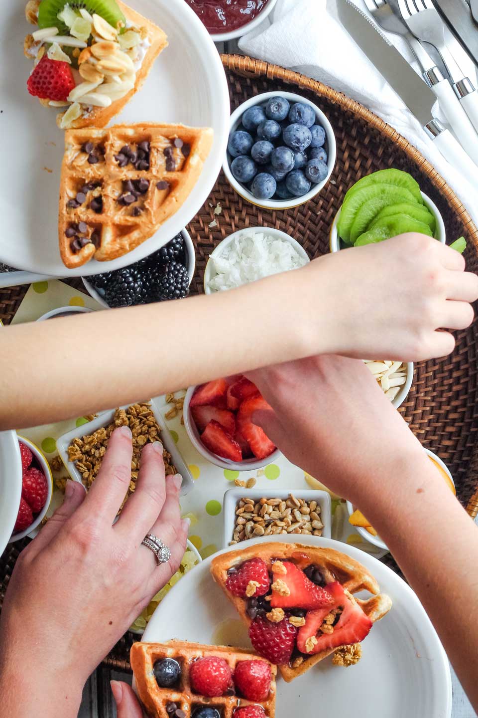 Overhead photo of three hands reaching across each other to load up plates of Galentine's Day waffles with various toppings and sprinkles from a waffle bar.