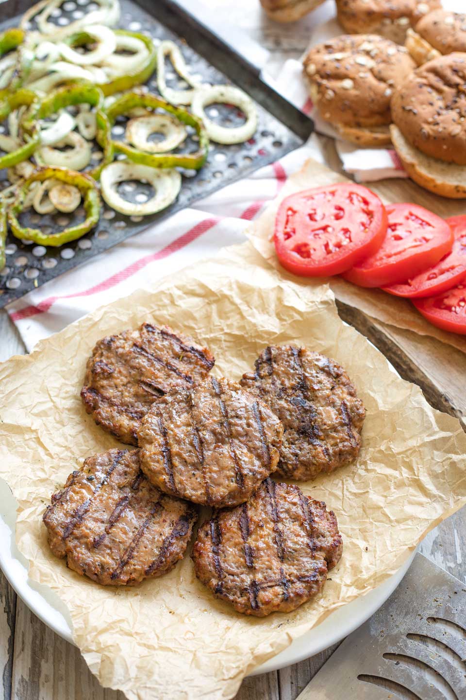 5 grilled burgers on a parchment-lined plate with hamburger buns, sliced tomatoes, grilled peppers and onions at the sides in background.